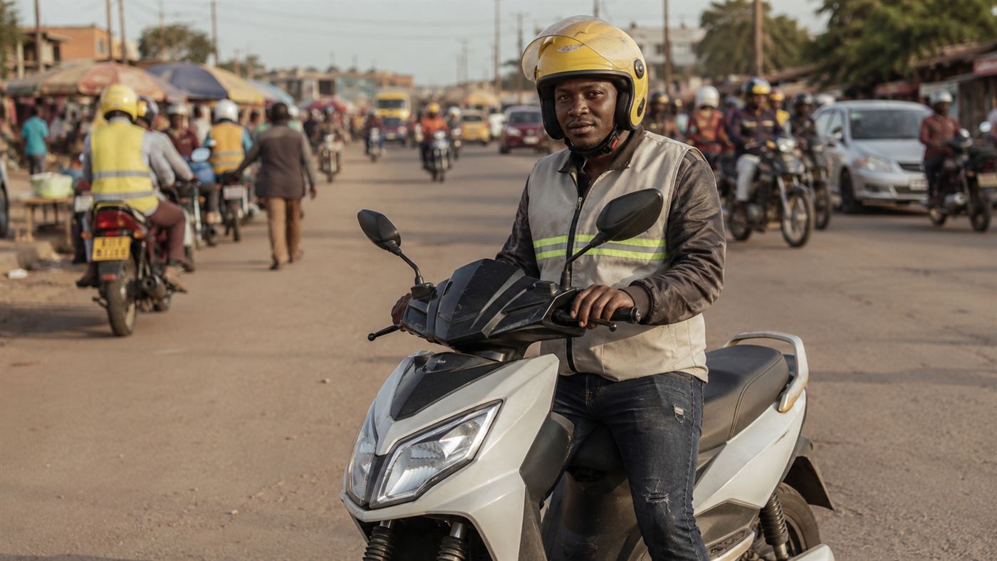 A motorcycle-taxi rider on an electric motorbike waiting in a busy African city street, with other riders and traffic behind