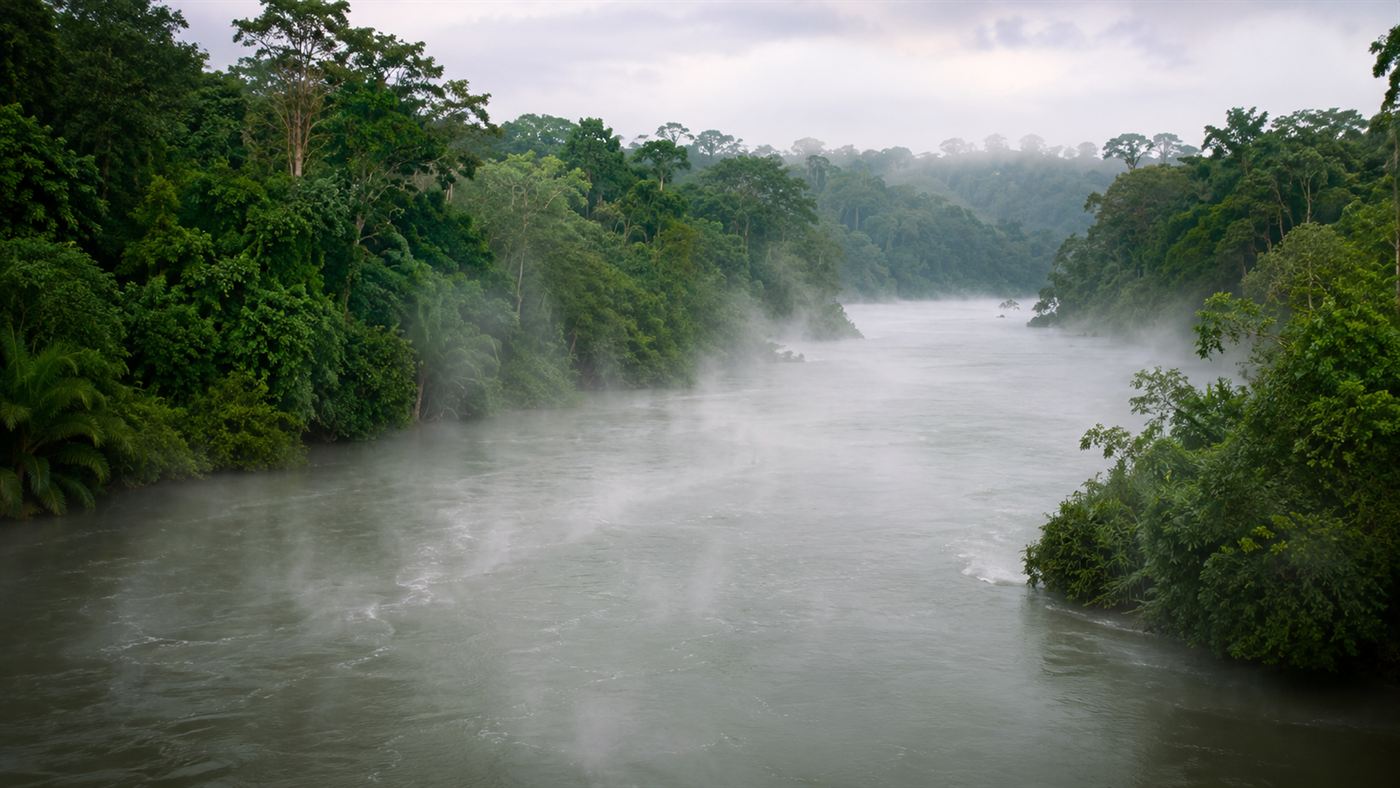 A wide river steaming heavily as it winds through dense green Amazon rainforest, with mist and vapour rising off the hot grey-green water