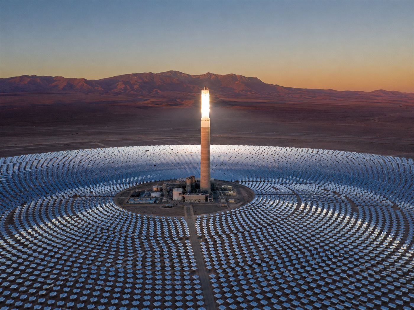 A tall glowing solar power tower surrounded by thousands of mirrors arranged in vast rings across the Atacama desert at dusk