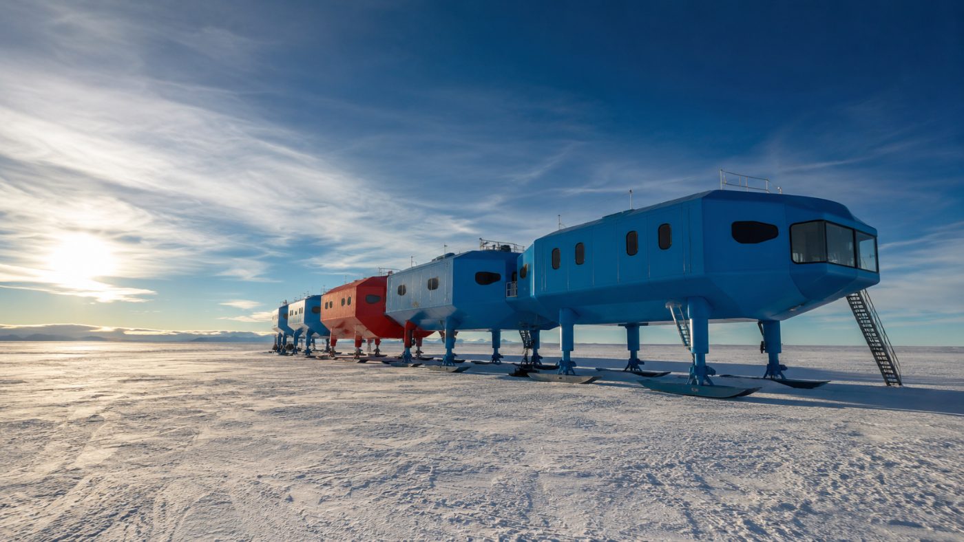 A row of blue and red modular pods on tall hydraulic legs and skis standing on the flat white Antarctic ice shelf under a dramatic polar sky