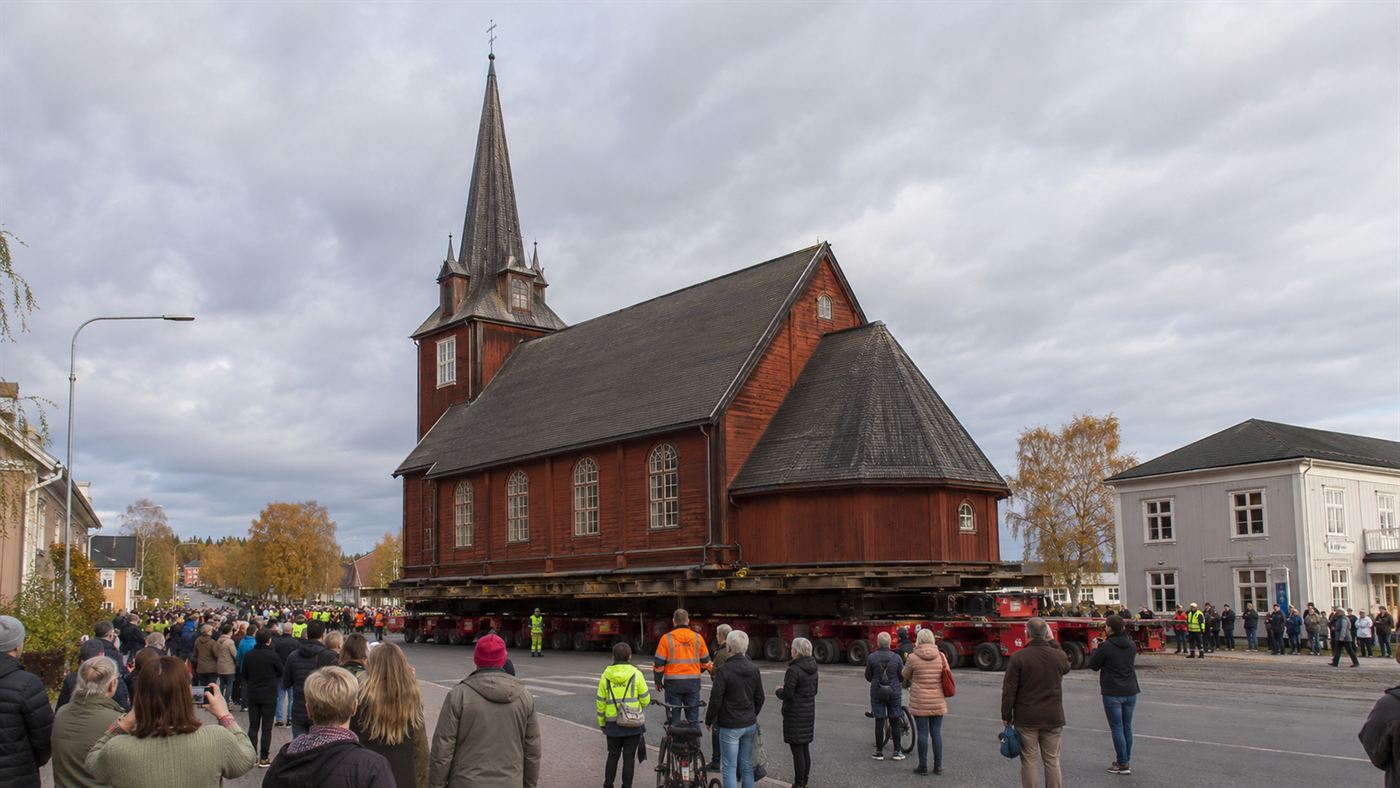 A large dark-red wooden church being transported whole on giant remote-controlled trailers along a road through an Arctic Swedish town, with crowds watching