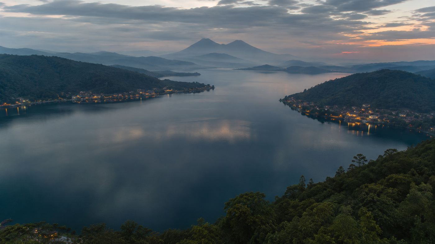 A calm, wide African lake at dusk ringed by green volcanic hills and lakeside towns, beautiful but still and ominous