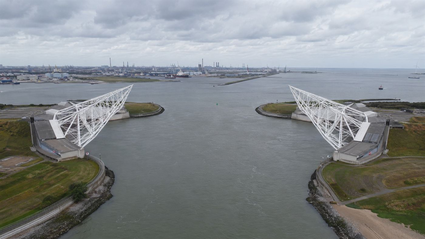 Aerial view of two enormous white steel truss arms of the Maeslant storm surge barrier resting on either bank of a wide shipping canal near Rotterdam