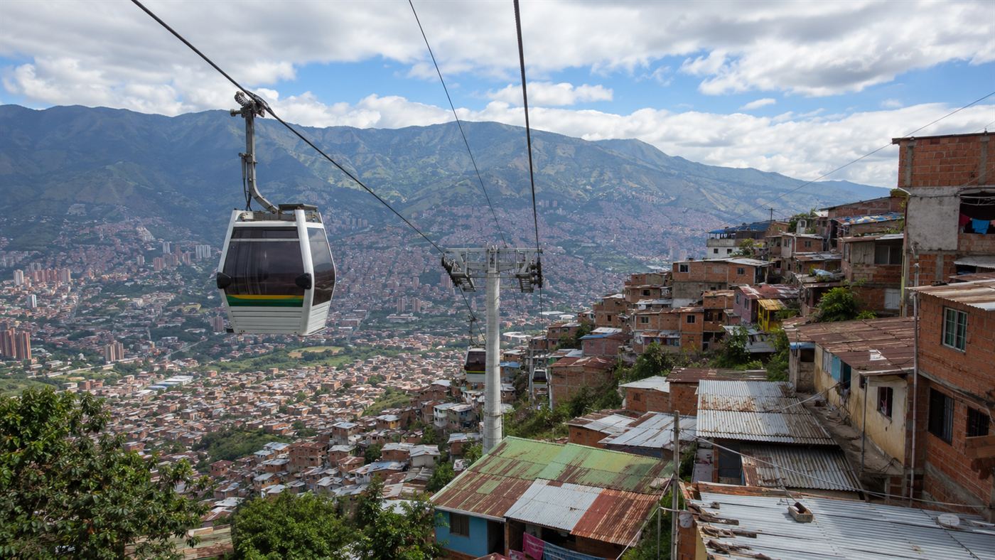 A bright cable car gondola rising on a steel cable over a dense hillside barrio of stacked brick houses in Medellin, with green mountains and the city below