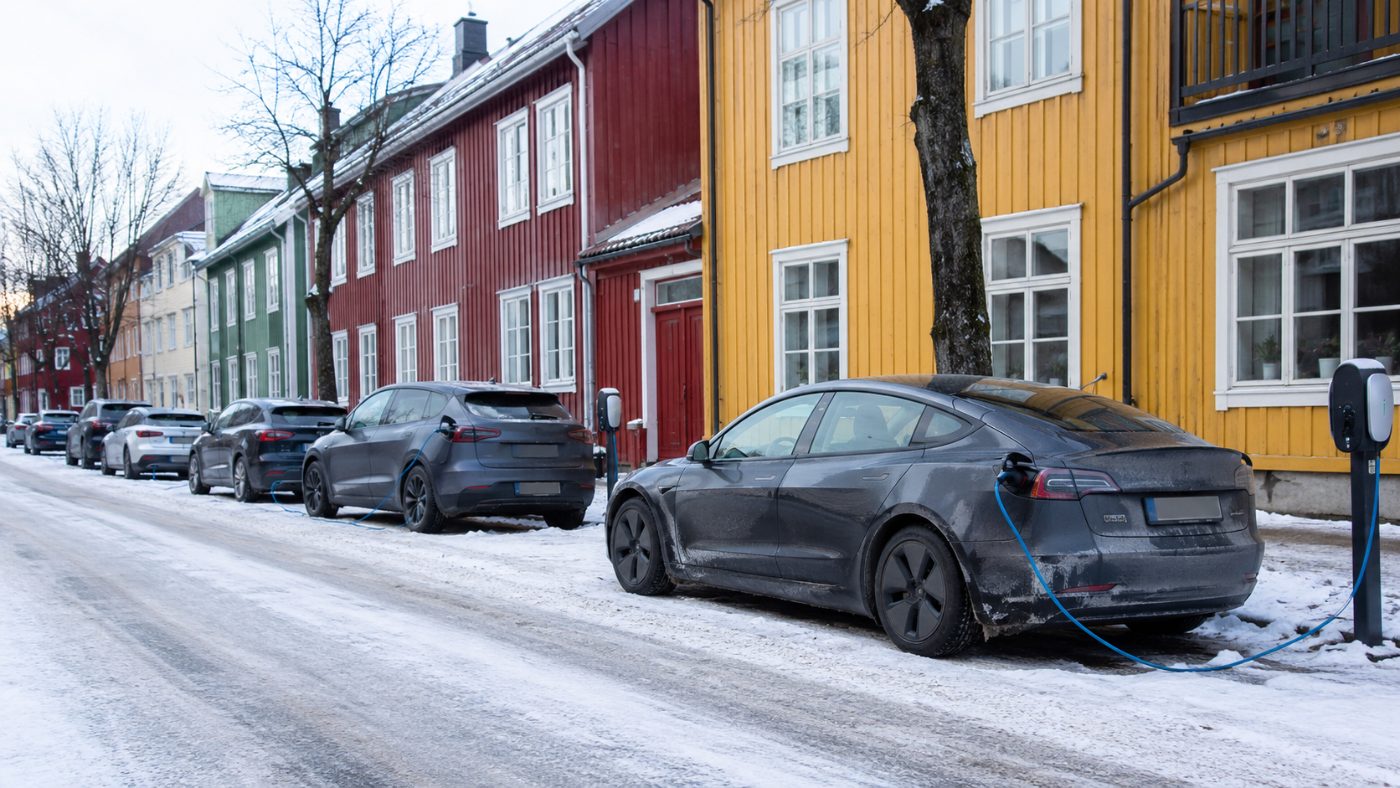 A snowy street in a Norwegian city lined with modern electric cars plugged into charging points, colourful wooden buildings behind them