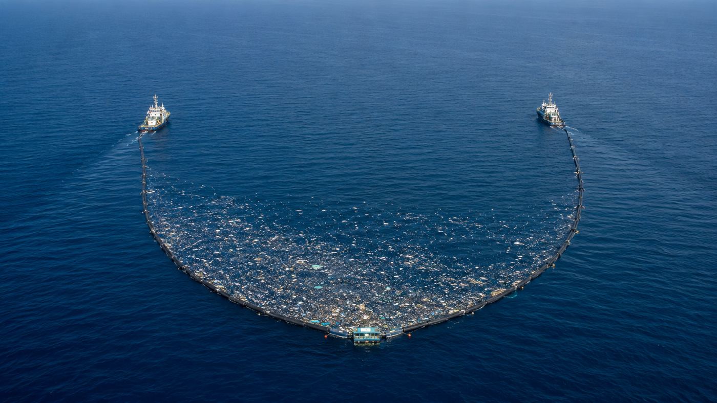 A long U-shaped floating barrier towed slowly between two ships across the open ocean, gathering floating plastic into a retention zone