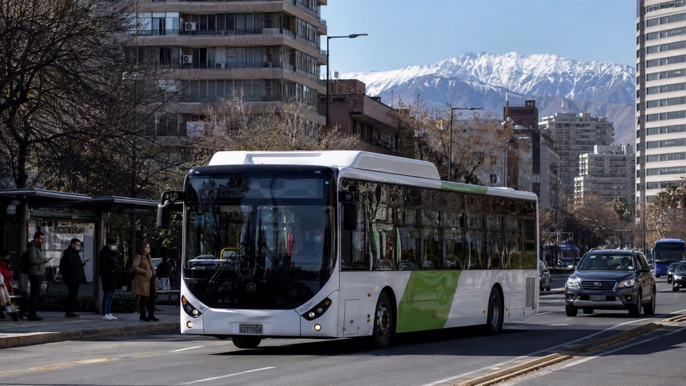 A modern white and green electric city bus on a Santiago street with the snow-capped Andes mountains rising behind the buildings