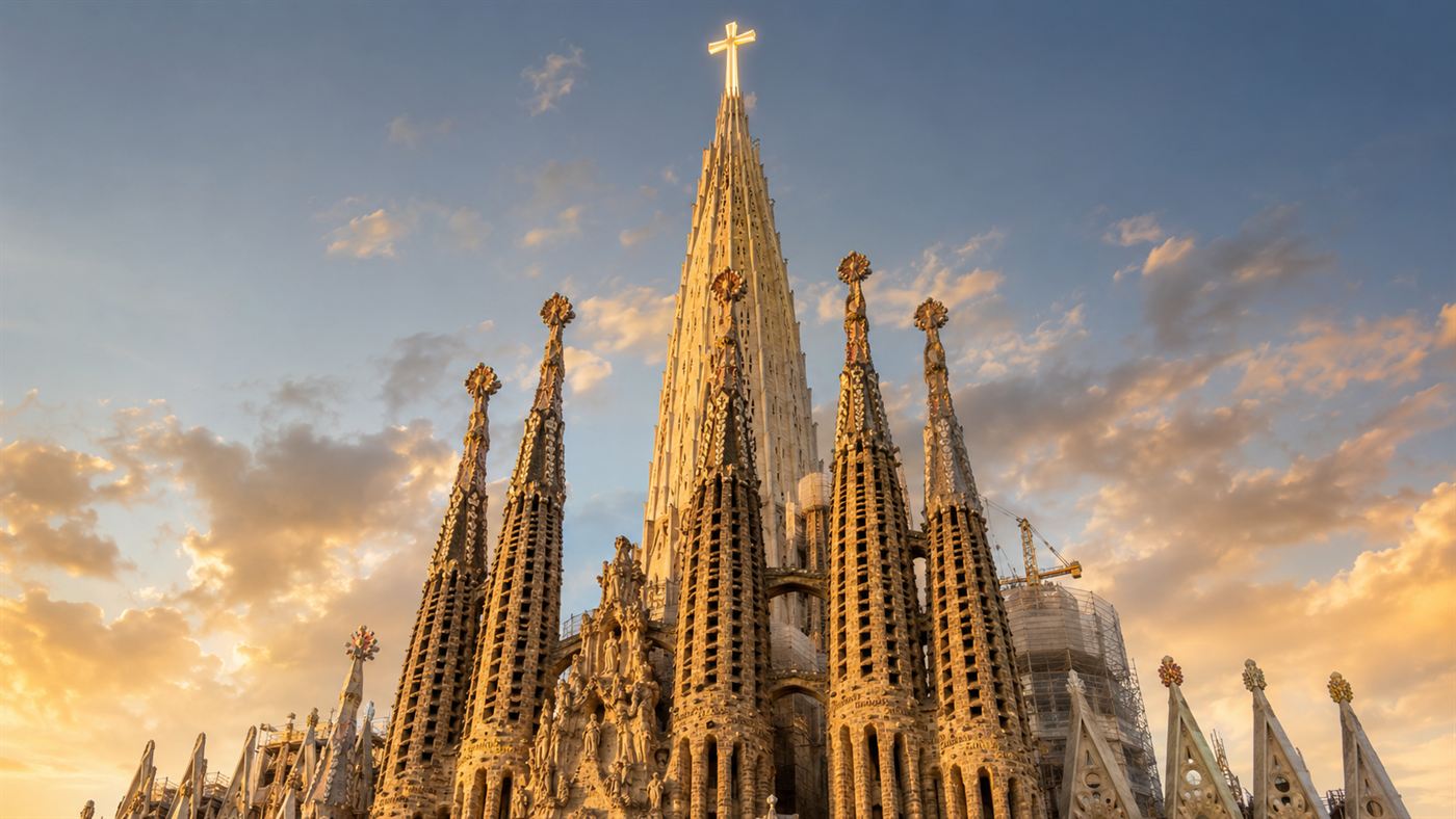 The Sagrada Familia basilica in Barcelona seen from below, its tall central tower crowned with a large cross rising above a cluster of stone spires in warm evening light
