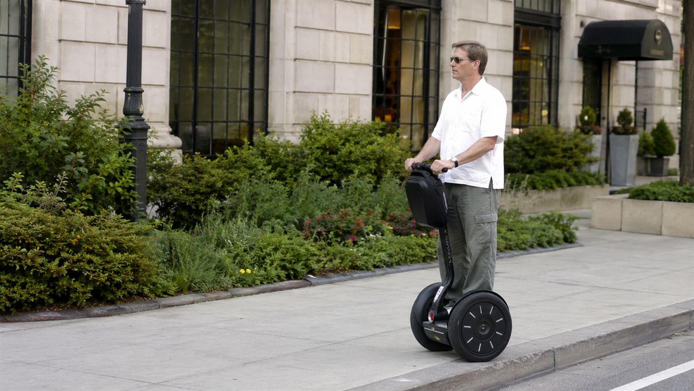 A person standing upright and riding a two-wheeled self-balancing Segway personal transporter along a city pavement