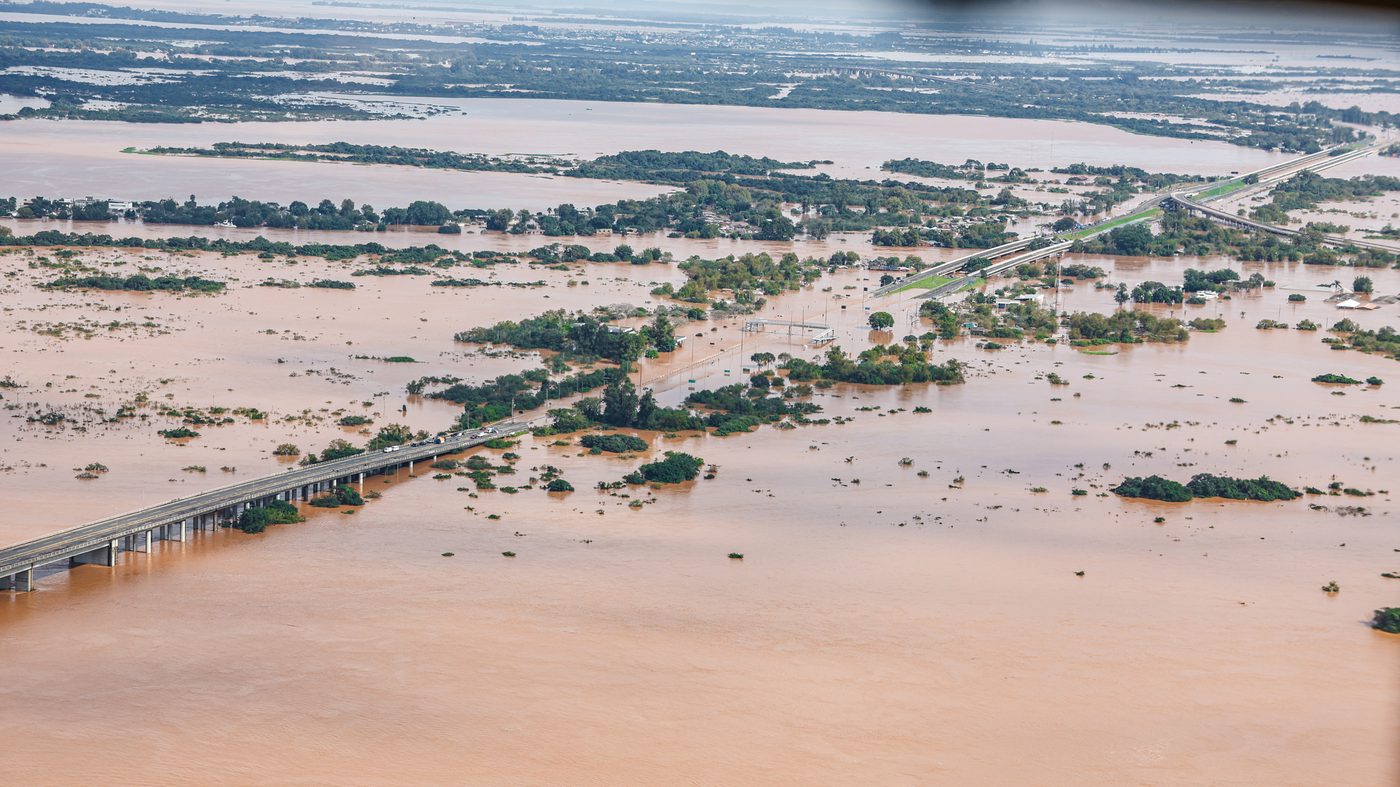 Vista aérea de uma rodovia submersa na Região Metropolitana de Porto Alegre (RS), com apenas o viaduto visível sobre a água marrom da enchente