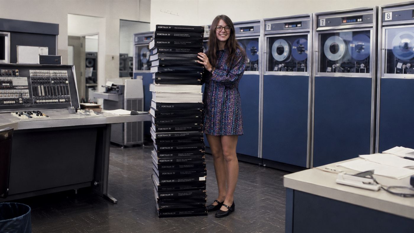 A woman scientist in the 1960s standing beside a stack of printed Apollo source code as tall as she is, evoking Margaret Hamilton