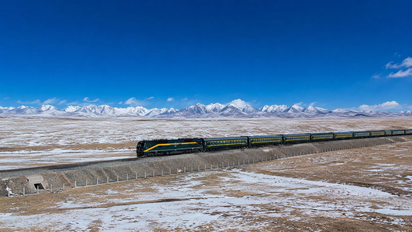 A train on the Qinghai-Tibet Railway crossing a vast high plateau with snow-capped mountains, the track raised on an embankment