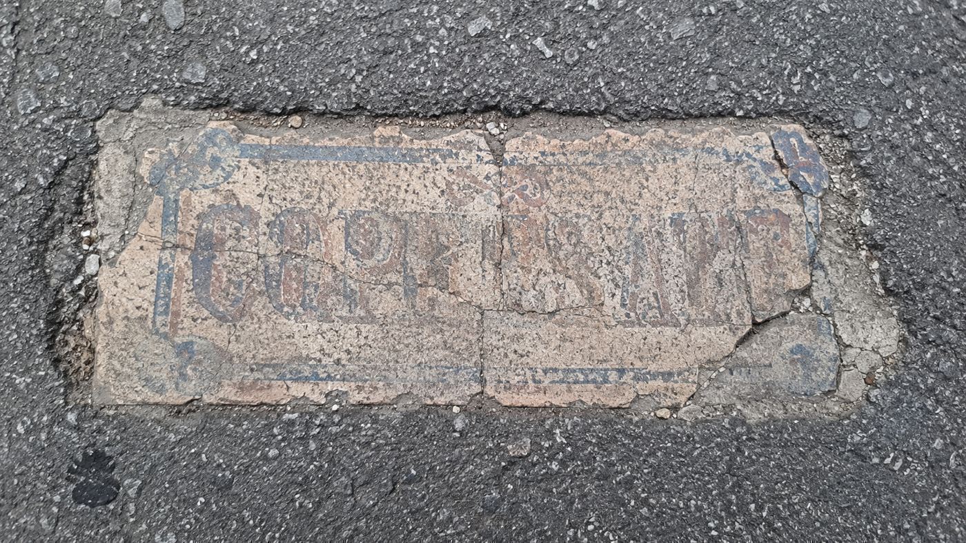 A worn handmade tile with faded lettering embedded flush in the cracked asphalt of a city street, a Toynbee tile
