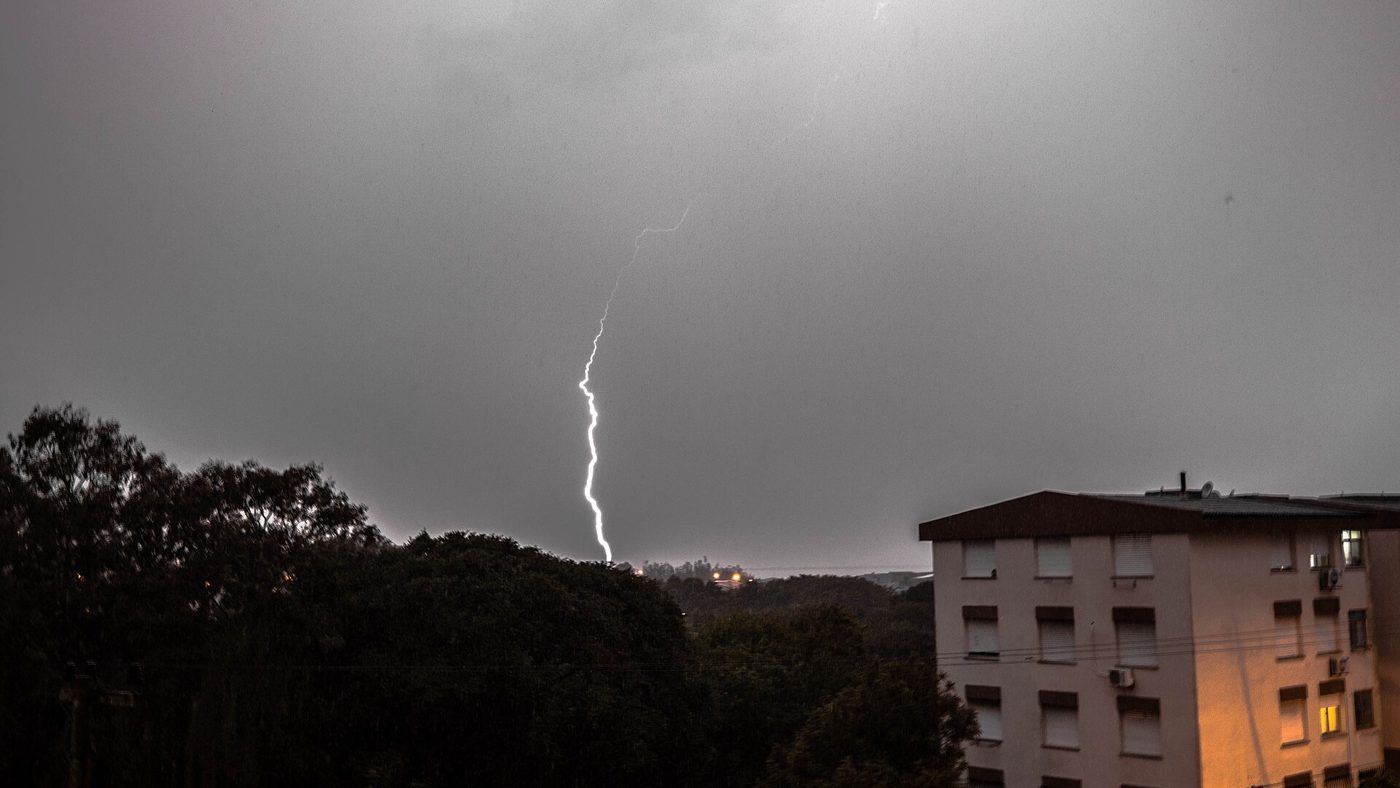 Raio desce de um céu cinzento sobre morro com árvores ao lado de prédio residencial em Rio Grande durante temporal
