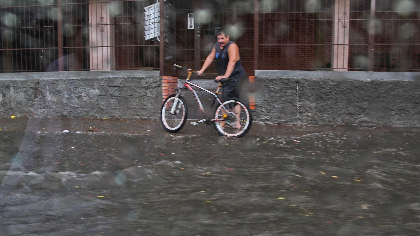 Homem empurra bicicleta por rua alagada durante forte temporal em Porto Alegre, com gotas de chuva na lente