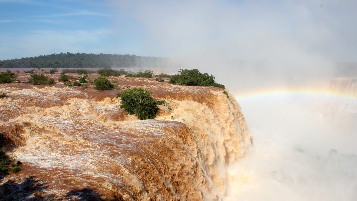 Água marrom e barrenta despencando nas Cataratas do Iguaçu em dia de cheia, com arco-íris na névoa à direita