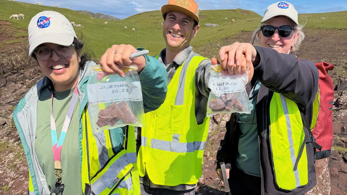 Três cientistas de colete amarelo mostram sacos com amostras de rocha na Baía de Clachtoll, na Escócia