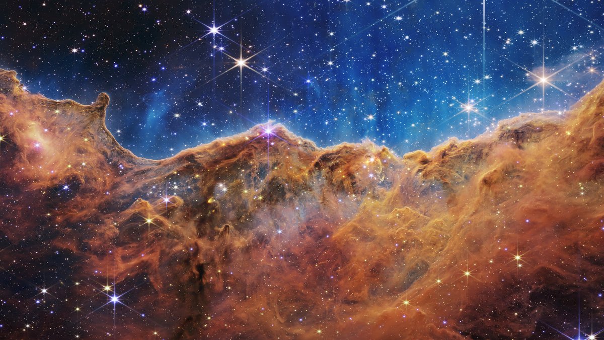 Paredões de gás e poeira alaranjados sob um céu azul cheio de estrelas, a borda da região NGC 3324 na Nebulosa de Carina vista pelo Webb