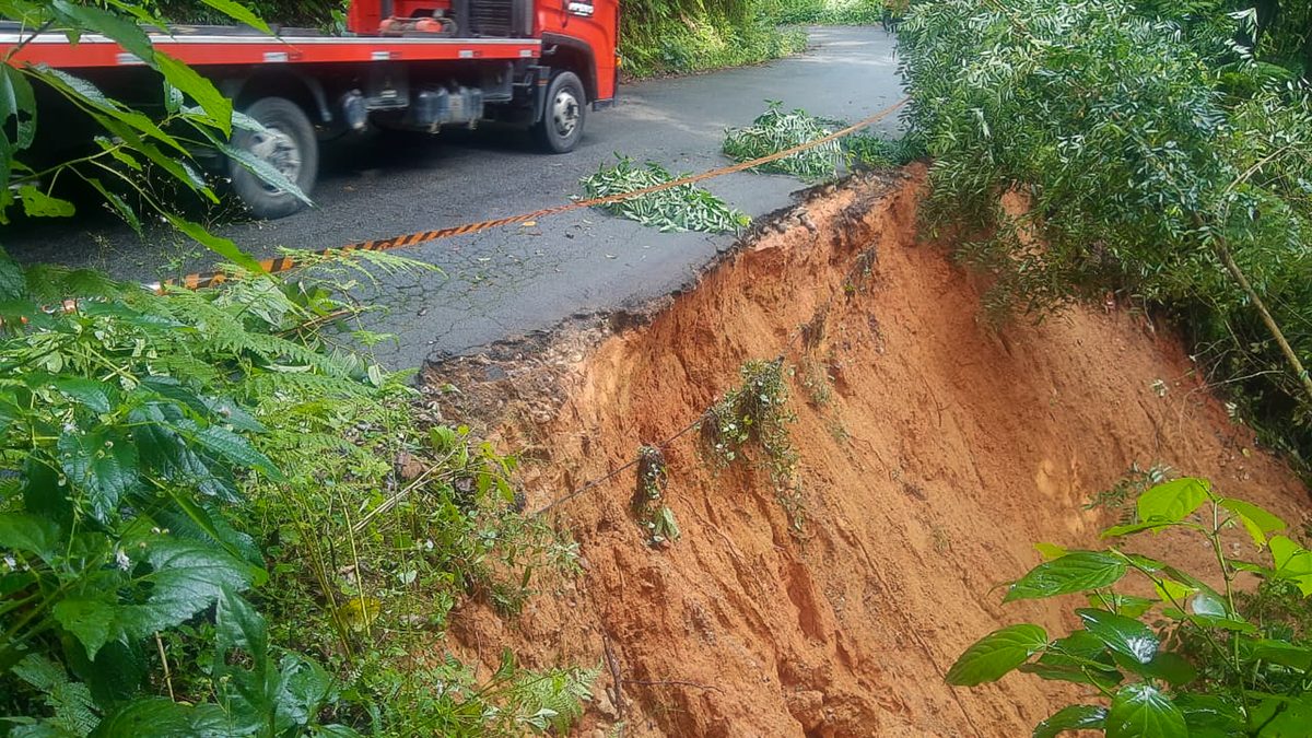 Trecho de estrada de asfalto cortado por um deslizamento, com a terra vermelha exposta, fita de isolamento e um caminhão guincho ao fundo