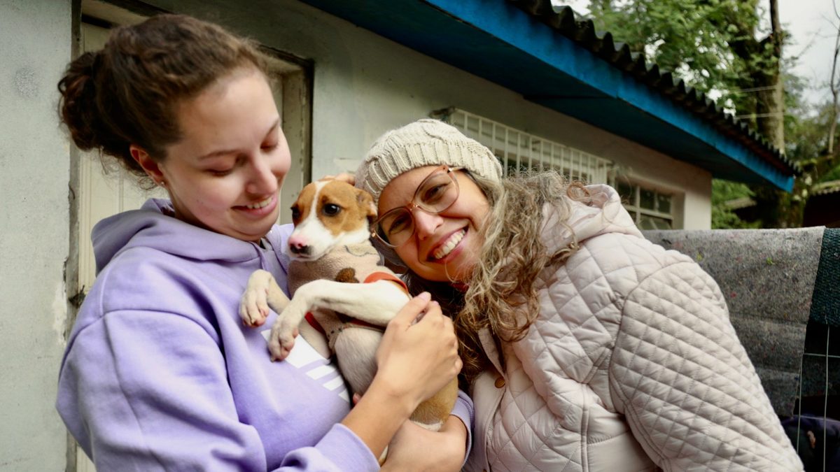 Duas mulheres sorrindo diante de uma casinha de cachorros, uma delas segurando um filhote branco e marrom no colo