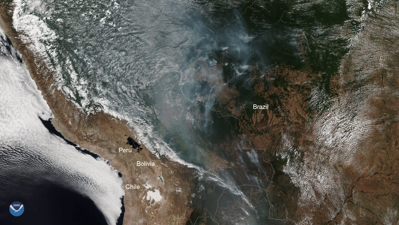 Imagem de satélite da América do Sul com os Andes, nuvens brancas e uma faixa de fumaça cinza sobre o Brasil e a Bolívia, com nomes de países