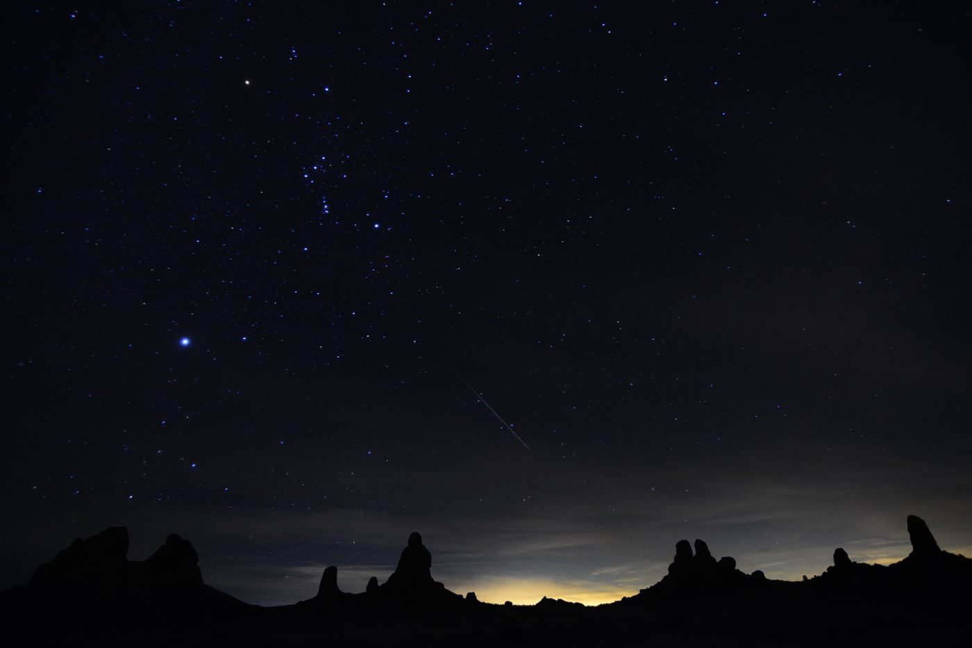 Céu estrelado com um meteoro riscando o alto, acima da silhueta de formações rochosas no deserto durante a noite.