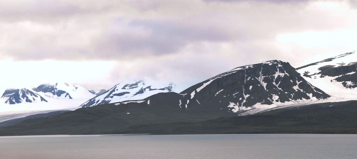 Montanhas escuras com neve e línguas de gelo do lado oeste de um fiorde de Svalbard, sob céu nublado no verão