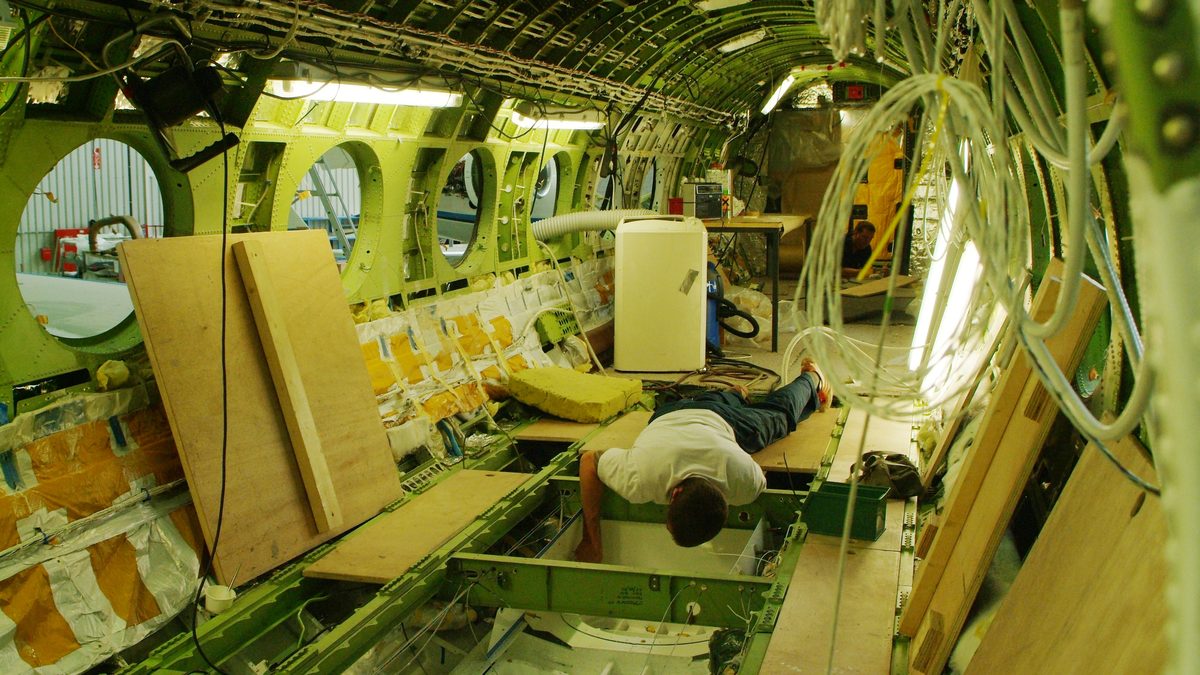 Interior da cabine do avião de pesquisa HALO sem acabamento, com fios soltos e um técnico trabalhando no piso aberto durante a conversão