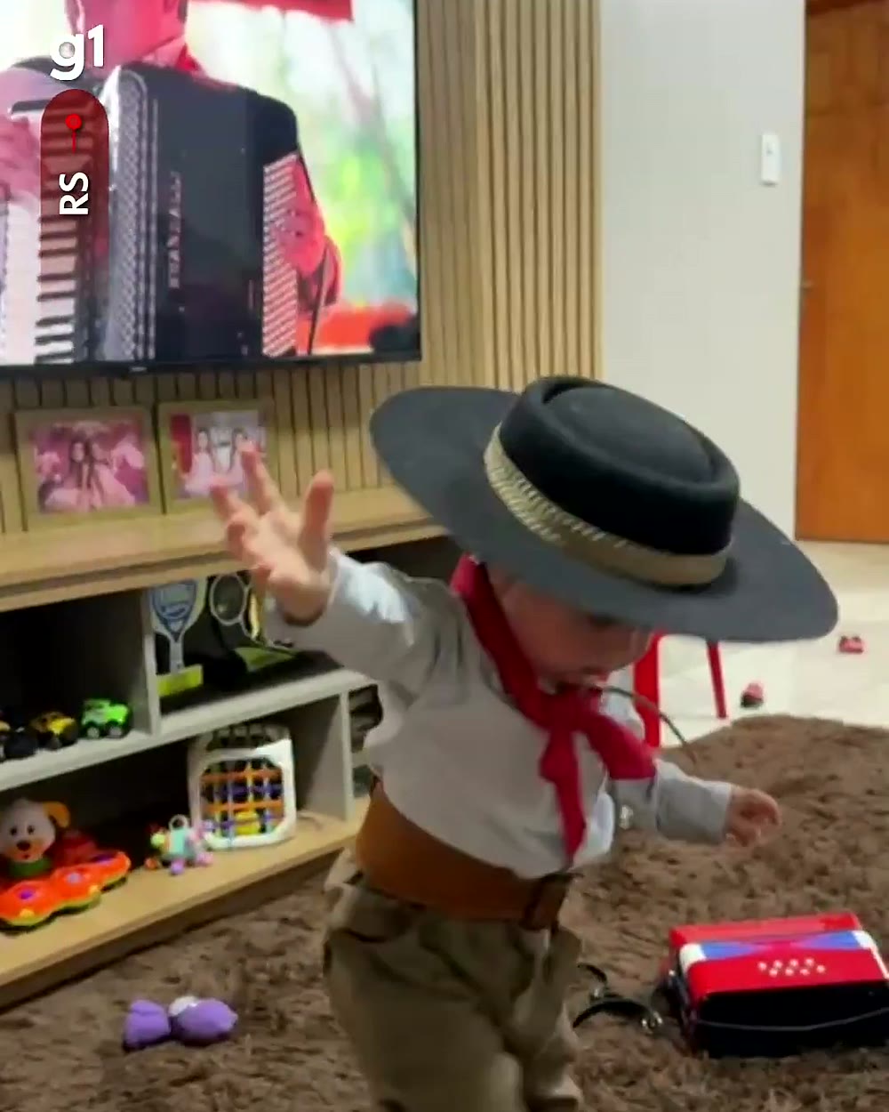 Joaquim de braços abertos dançando música gaúcha na sala, com o sanfoneiro na TV ao fundo