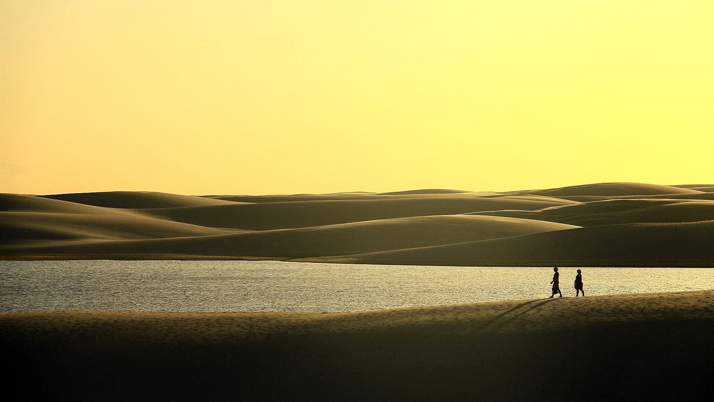 Duas pessoas caminham por uma duna dos Lençóis Maranhenses ao entardecer, ao lado de uma lagoa entre as dunas.