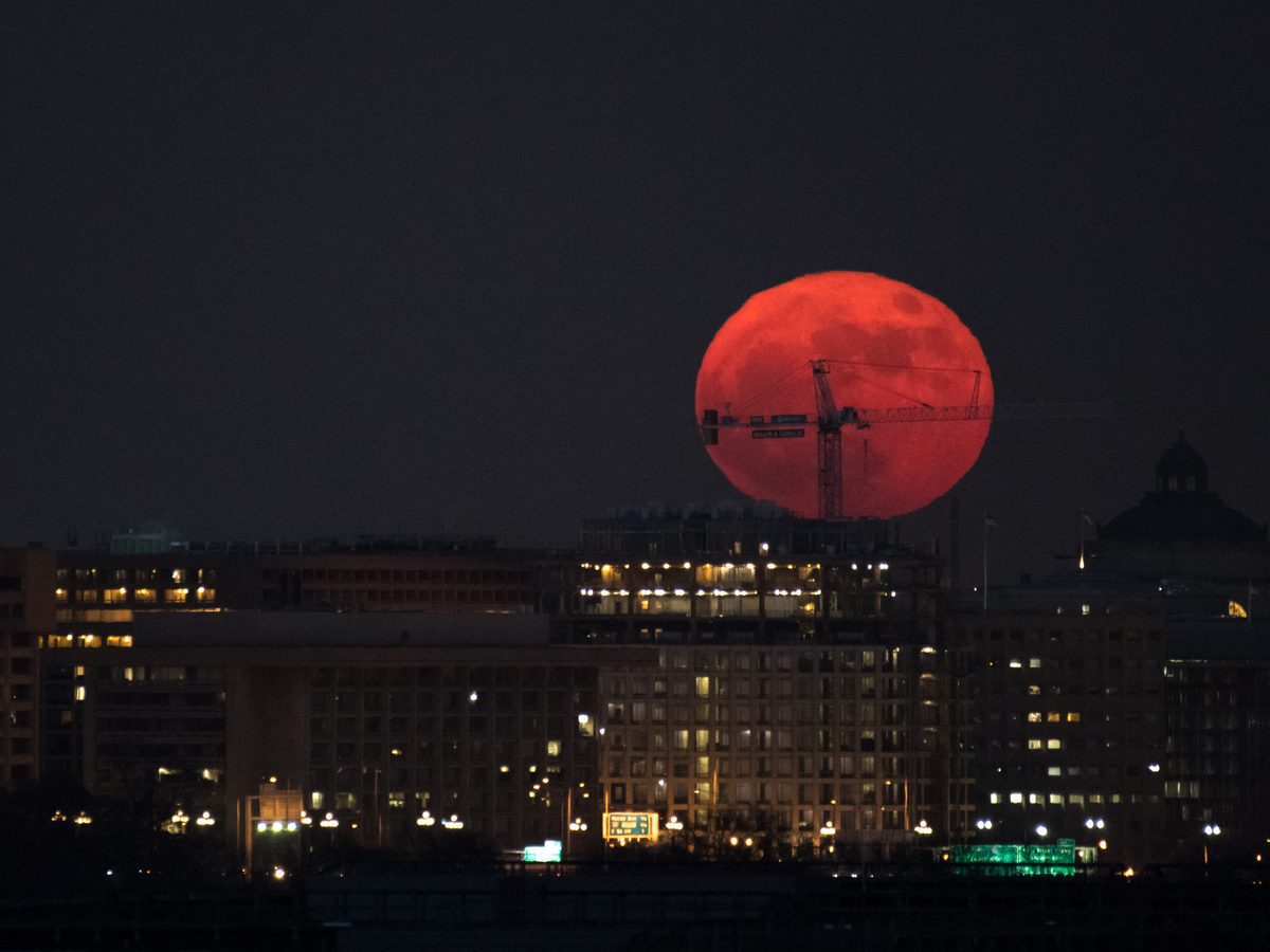 Lua cheia enorme e avermelhada atrás de prédios iluminados e de um guindaste, fotografada com lente de longo alcance