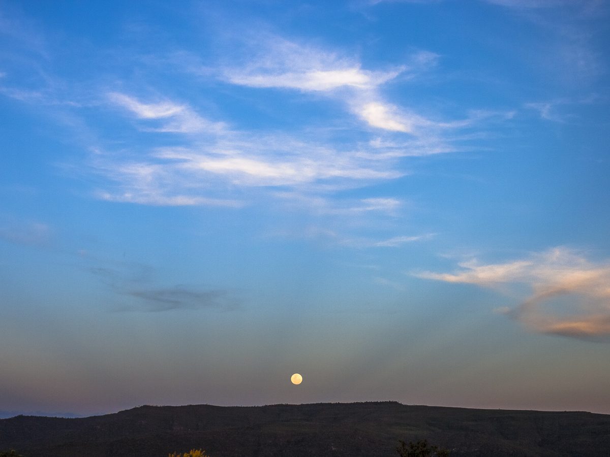 Lua cheia pequena logo acima de uma serra escura, com céu azul e nuvens finas, vista sem zoom