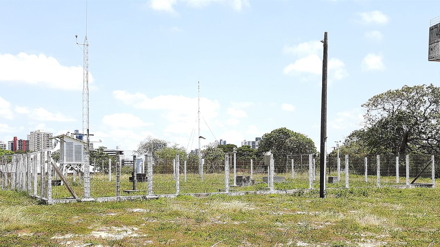 Estação meteorológica do Inmet cercada por tela em Natal, com abrigo de instrumentos, torre de vento e pluviômetros