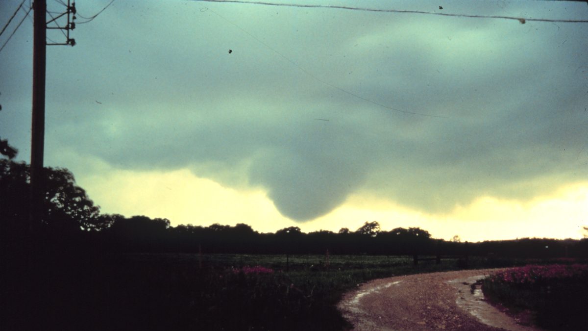 Nuvem em forma de funil descendo de um céu esverdeado escuro sobre campo e estrada de terra, ao lado de um poste