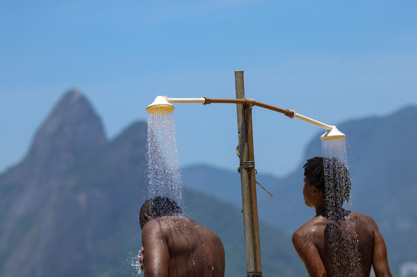 Dois banhistas de costas se refrescam sob um chuveiro de praia no Rio de Janeiro, com o morro Dois Irmãos ao fundo