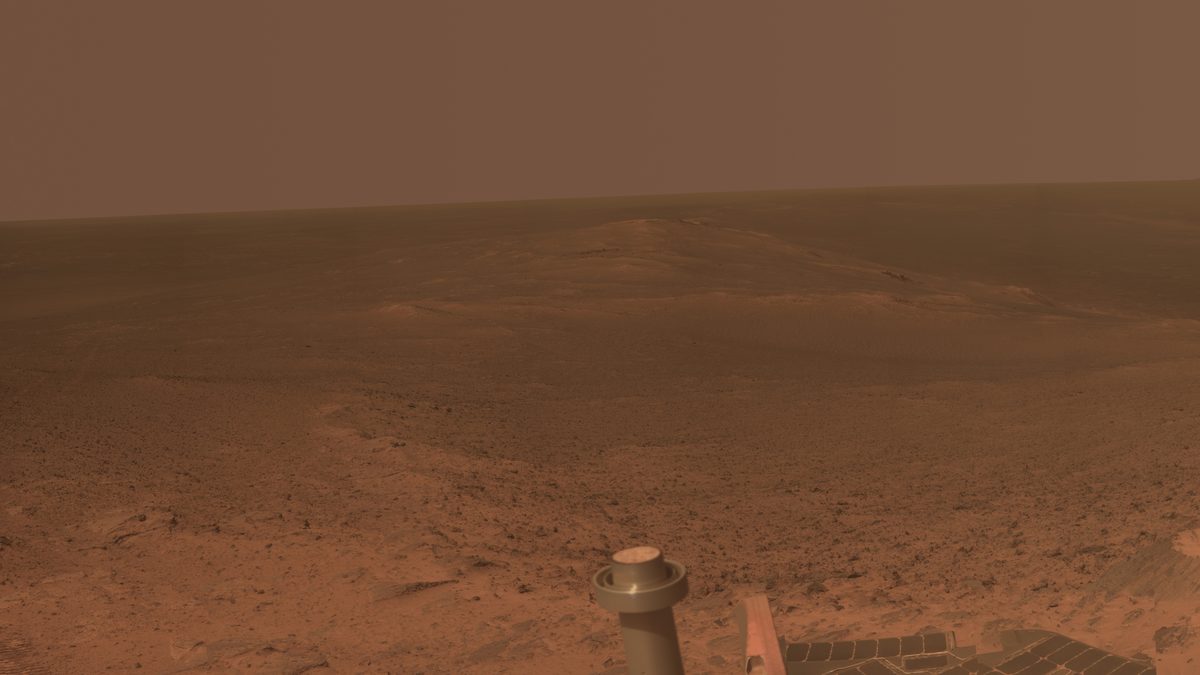 Planície avermelhada de Marte vista do topo de uma borda de cratera, com o mastro e o painel solar do rover Opportunity em primeiro plano