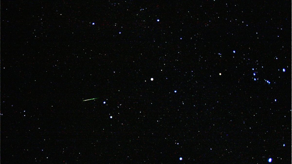 Céu escuro cheio de estrelas com um risco verde de meteoro Orionídea à esquerda e a constelação de Órion no lado direito
