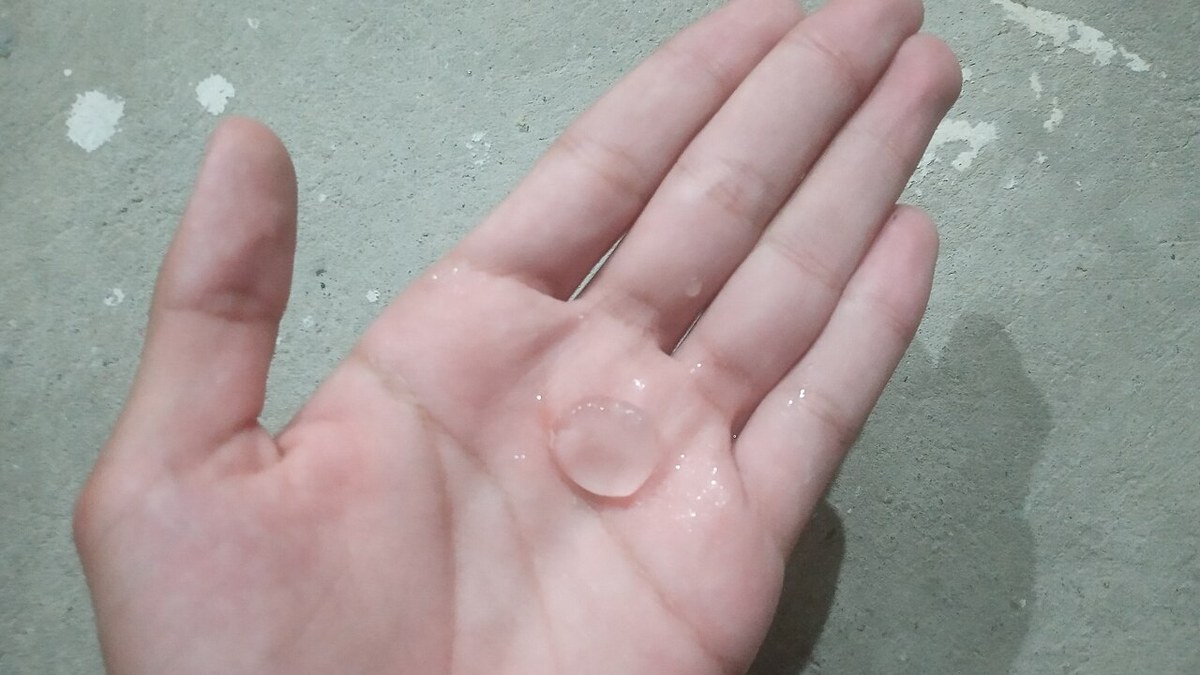 Mão aberta segurando uma pedra de granizo transparente do tamanho de uma moeda, sobre piso de concreto cinza