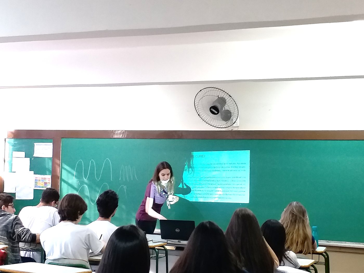 Professora apresenta aula de química com projetor na lousa verde para alunos do ensino médio sentados em carteiras, com ventilador na parede