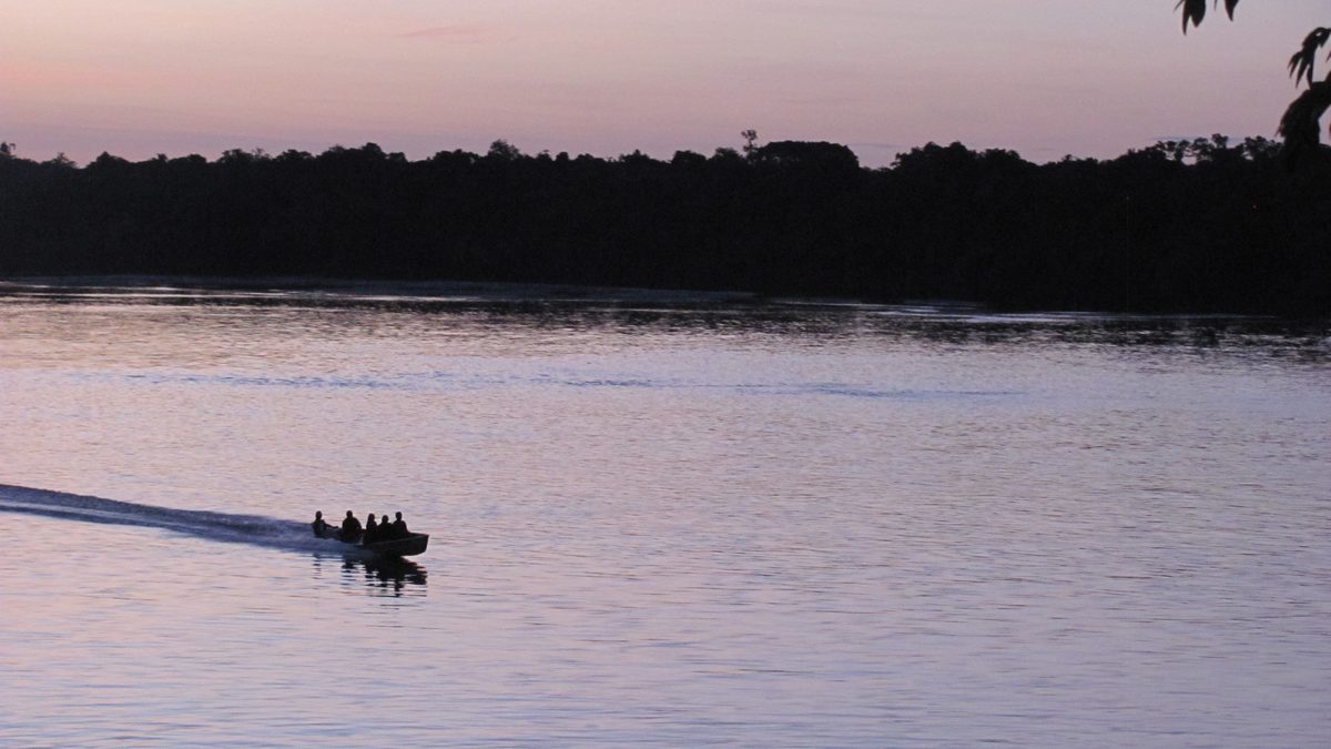 Barco pequeno com pessoas cruzando a água calma do rio Inírida ao entardecer, com a floresta escura ao fundo