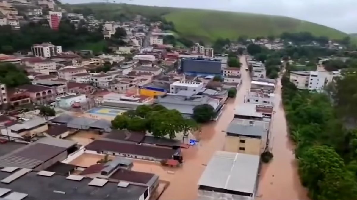 Vista aérea de uma rua alagada por água marrom entre prédios e telhados, com morros verdes ao fundo, na Zona da Mata mineira