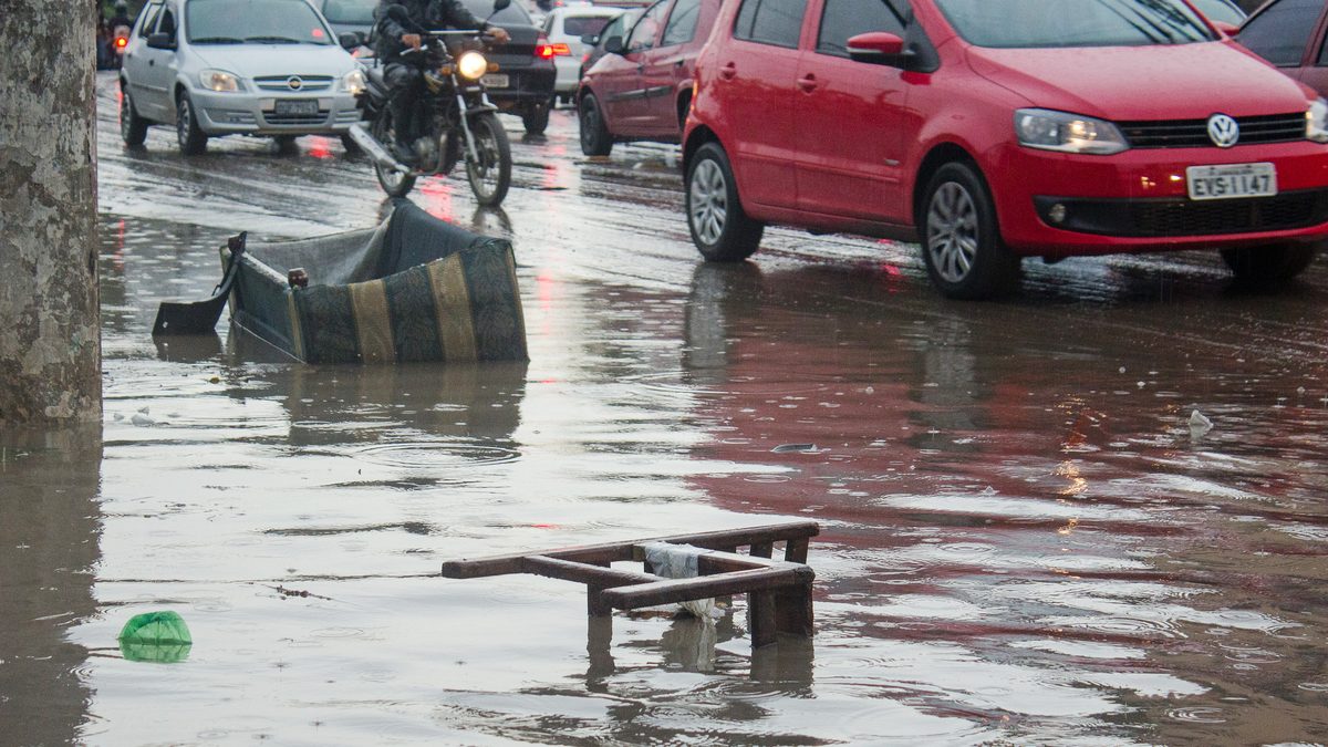 Rua de Taboão da Serra com água e objetos no asfalto, motociclista e carros passando devagar depois da chuva, um caso de alagamento urbano