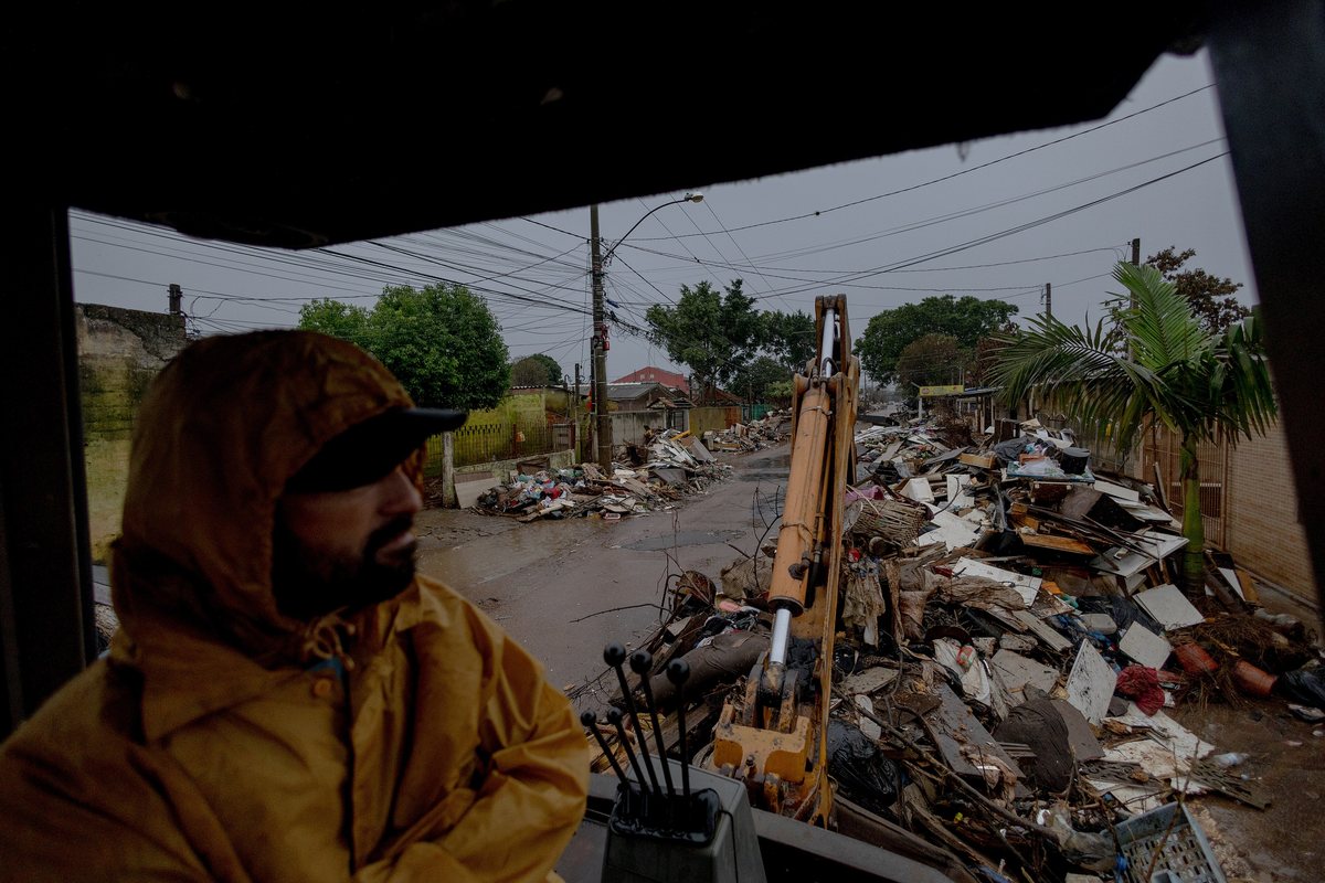 Rua tomada por montanhas de entulho e móveis estragados depois da enchente, com uma retroescavadeira e um homem de capa de chuva