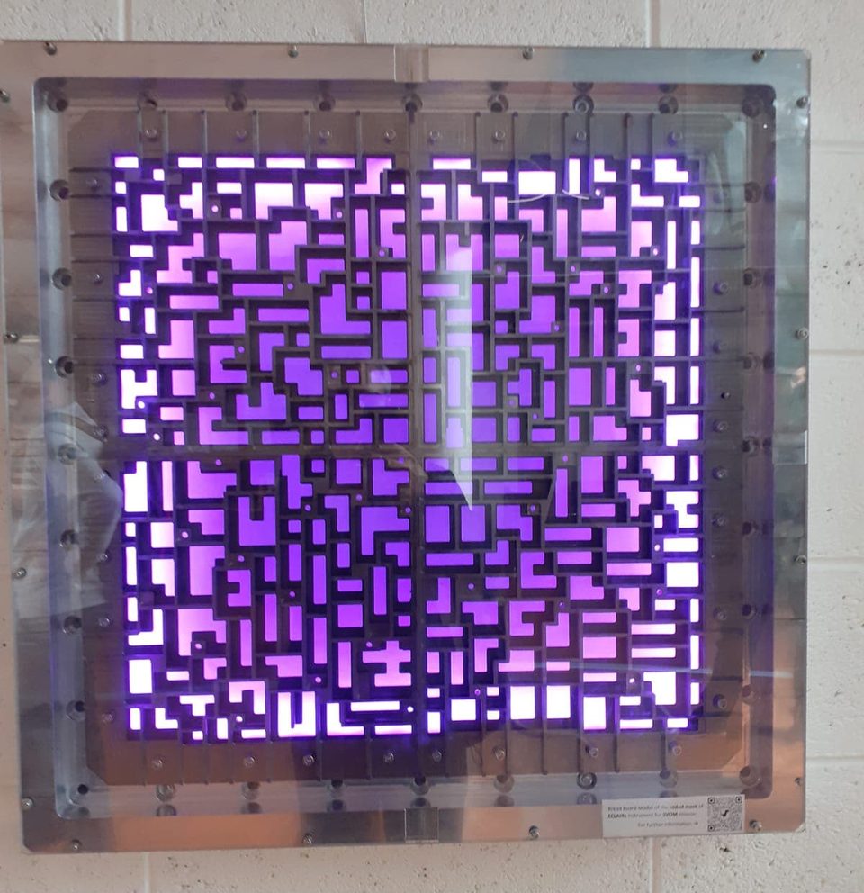 Placa metálica quadrada cheia de recortes irregulares, iluminada por trás em tom roxo, protótipo da máscara de um detector de raios X