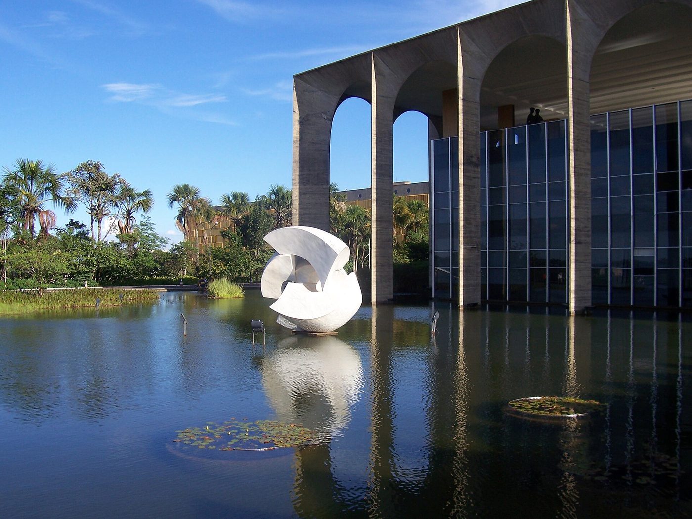 Escultura branca Meteoro no espelho d'água do Palácio do Itamaraty, com folhas flutuantes e palmeiras ao fundo