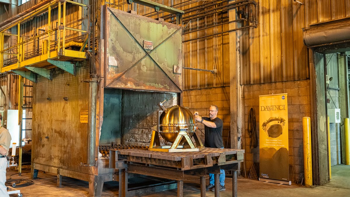 Técnico mexe na sonda DAVINCI de aço, sobre um suporte, diante da porta aberta do forno industrial, ao lado de um banner da missão